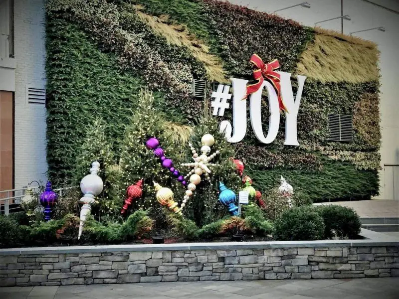 New Project (26) A vertical garden wall displays "#JOY" in large letters. Below, festive ornaments and small Christmas trees are arranged on a stone platform.