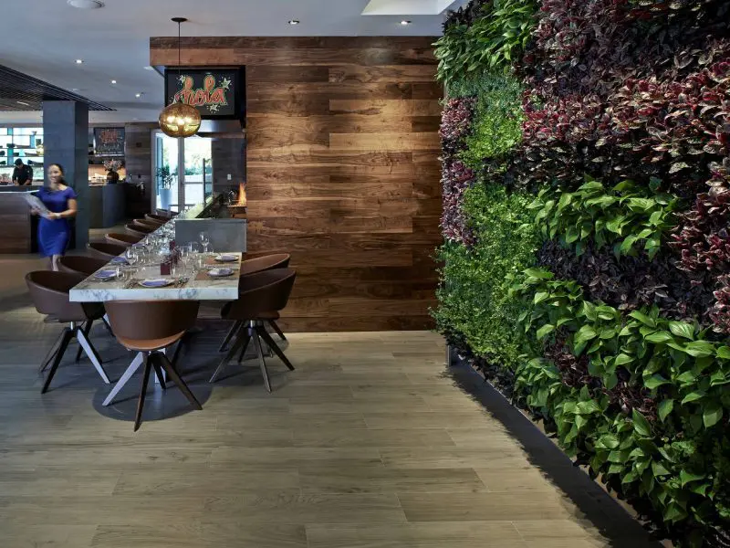 New Project (33) Modern restaurant interior with a wooden table and chairs, a green plant wall on the right, wooden paneling on the left, and warm lighting. A person is walking in the background.