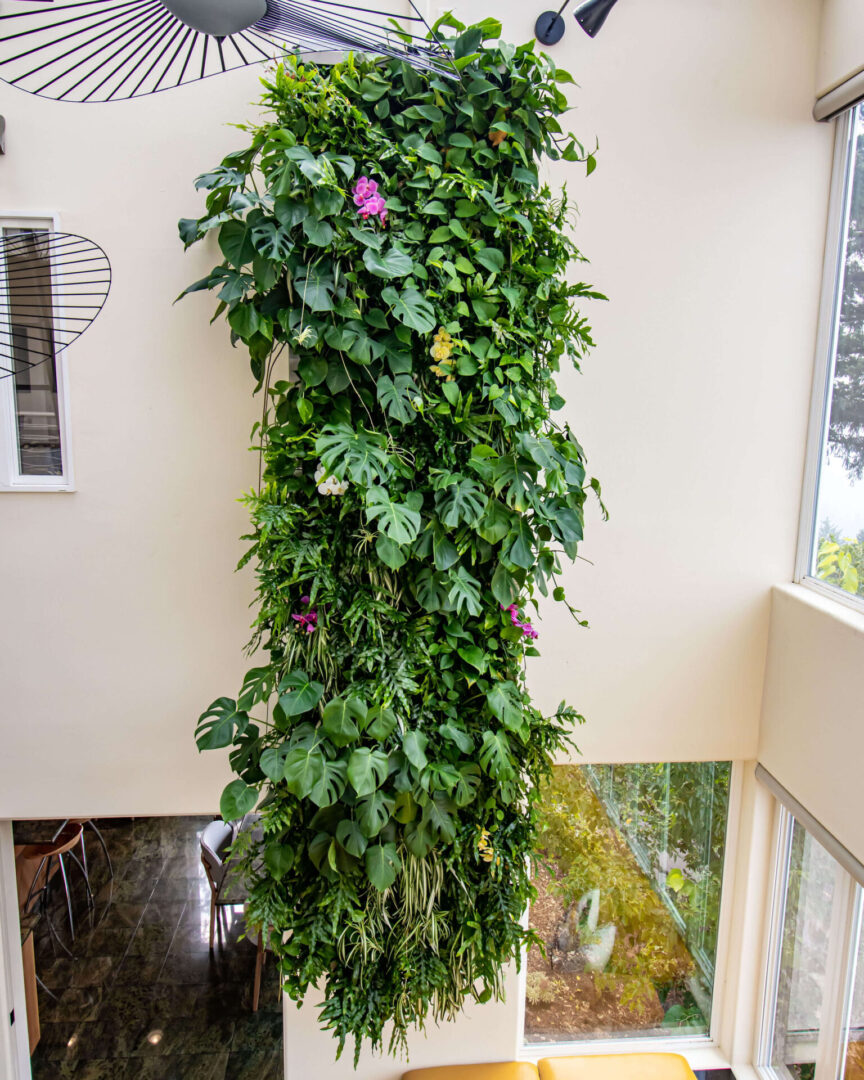 Copy of Petals_Frong-9264 Vertical indoor garden with lush green foliage and a few pink flowers, set against a white wall in a room with large windows and modern light fixtures.